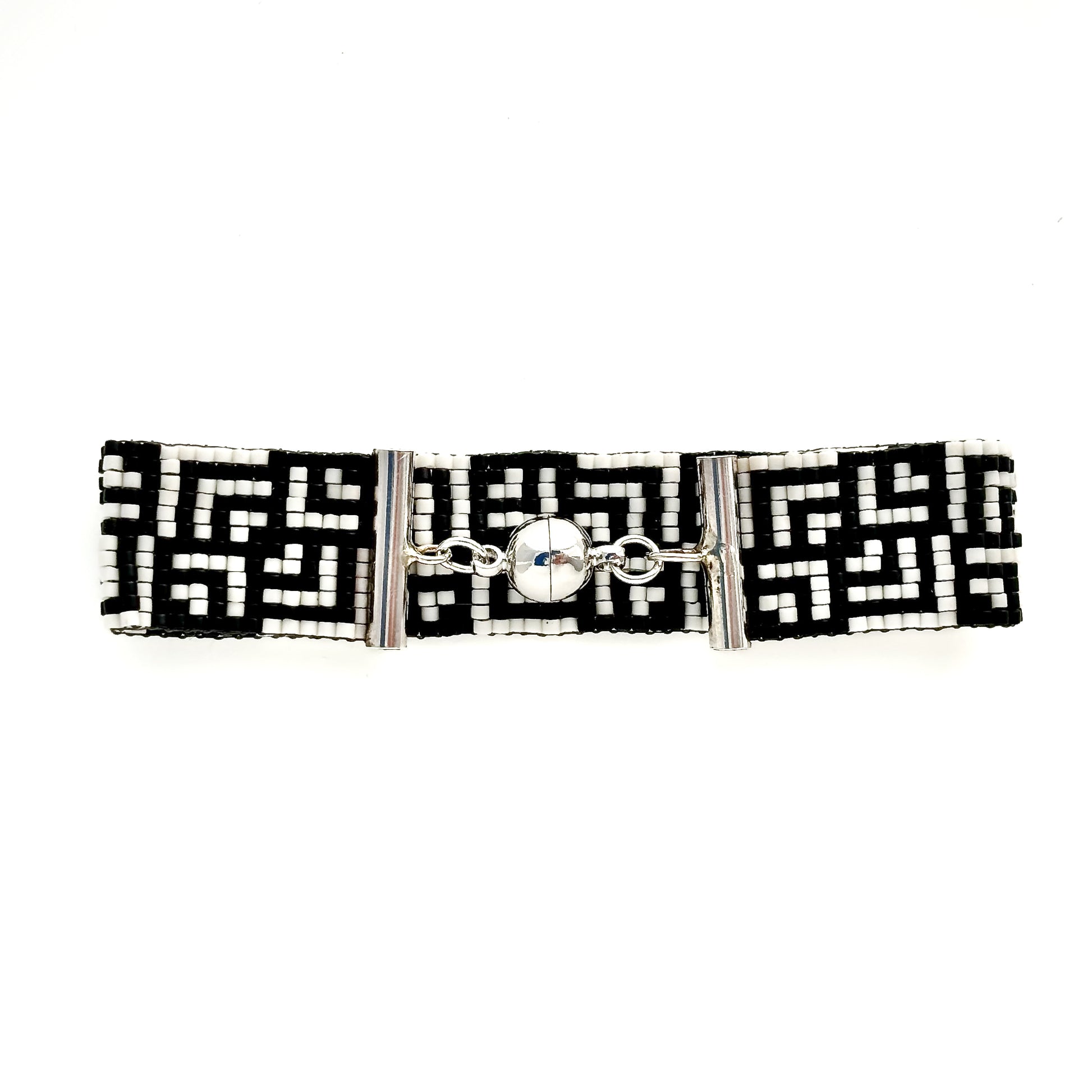A flat lay shows a beaded bracelet with a black and white Greek key pattern and a silver magnetic clasp.