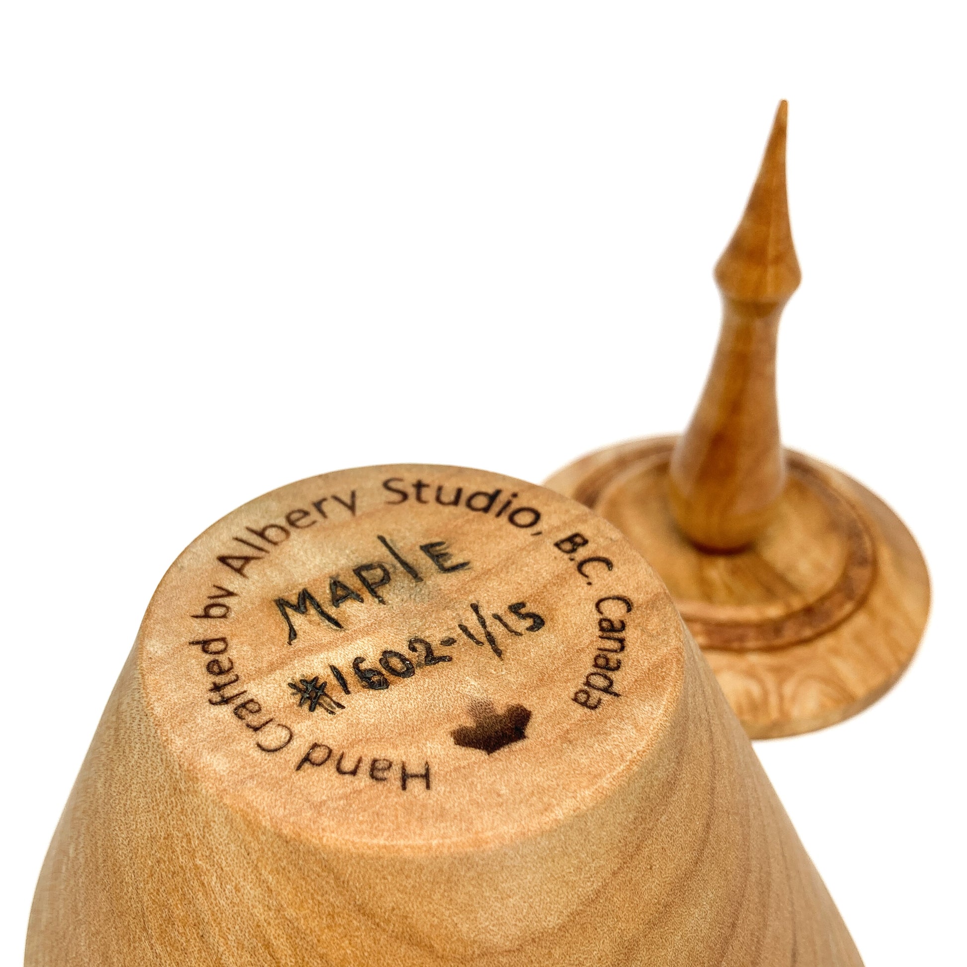 A turned wooden box lid with a pointed finial, showing the maker's mark on the bottom.