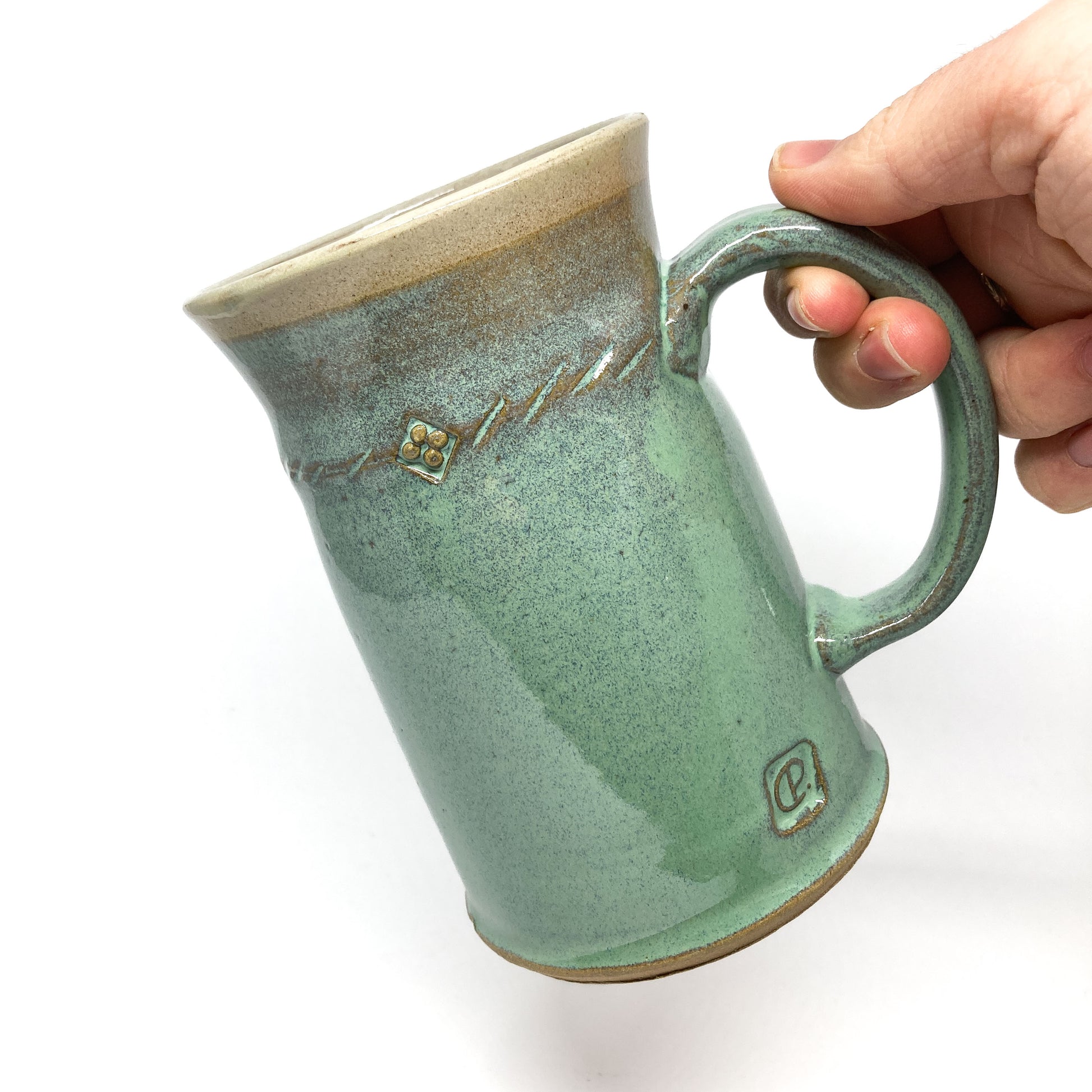 Hand holds a seafoam green glazed ceramic stein with a rope design and a maker's mark.