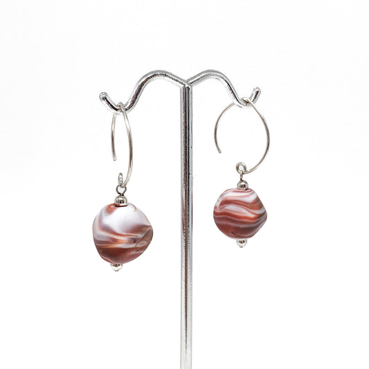 A pair of earrings with brown and white swirl glass beads hanging from silver hoops on a metal stand.