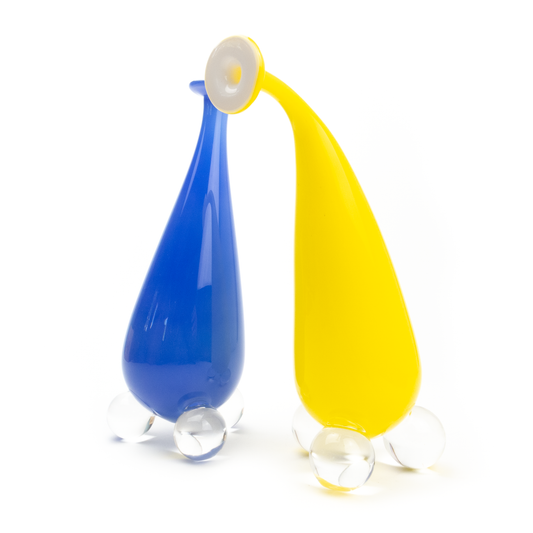 Two stylized glass figures, one blue and one yellow, with rounded bodies, curved necks, and resting on clear glass spheres.