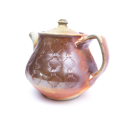 A wood-fired ceramic teapot with a lid and handle features impressed pomegranate designs on its surface.
