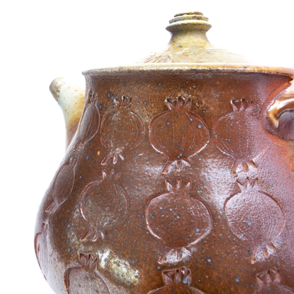 A close-up shows the textured surface and impressed pomegranate designs of a wood-fired ceramic teapot.