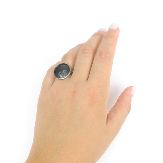 A hand wears a silver ring with a large, round, dark textured face featuring a radiating design.