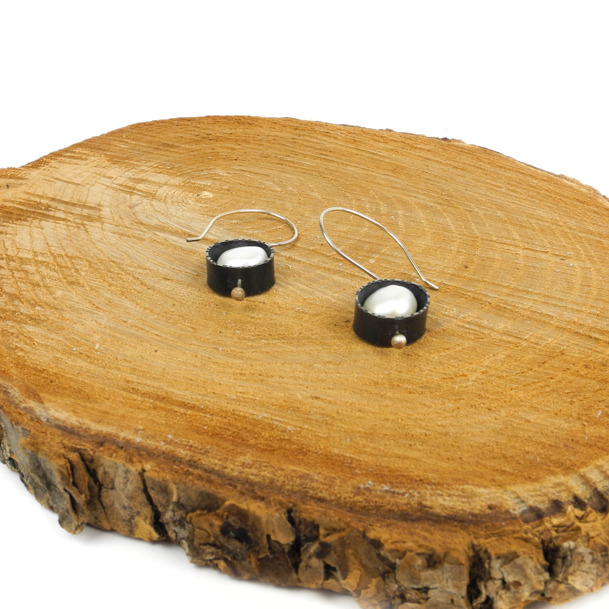 Two silver earrings with baroque pearls in textured circular settings rest on a rustic wooden slice.