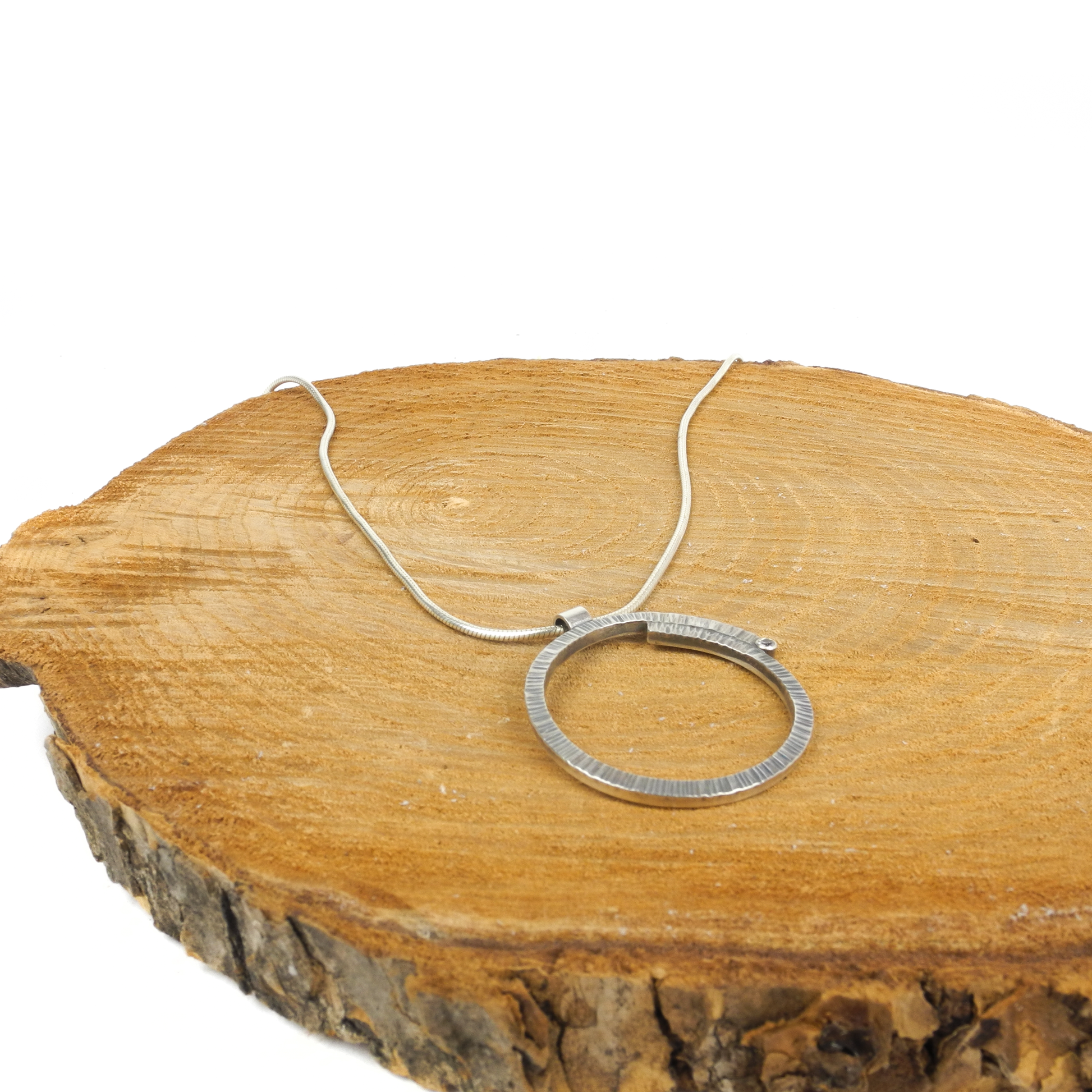 A metallic pendant with a textured circular design rests on a wooden surface, attached to a chain.